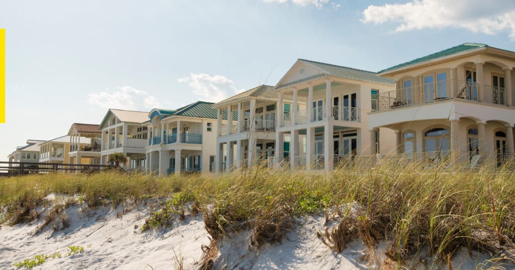 Tan coastal homes stand behind long grass beach vegetation and sandy beach